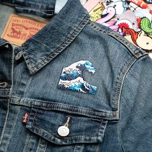 May include: A blue and white embroidered patch of the Great Wave off Kanagawa, a famous Japanese woodblock print by Hokusai, on a denim jacket.