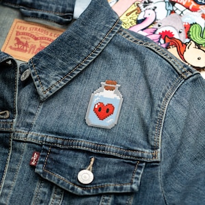May include: A pixelated patch featuring a potion bottle with a red heart inside. The bottle has a light blue liquid and a cork stopper. The patch is attached to a denim jacket, with a Levi Strauss & Co. tag visible.