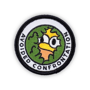 May include: A round green patch with a white border and black stitching. The patch has a cartoon face with a yellow head and black eyes. The text "AVOIDED CONFRONTATION" is written around the edge of the patch.