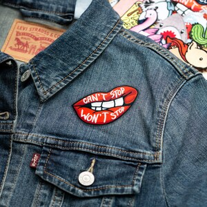 May include: A red embroidered patch with a pair of lips and the text "Can't Stop Won't Stop" sewn onto a blue denim jacket.