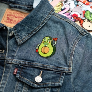 May include: A green avocado-shaped robot with red, blue, and yellow details is wearing a bow tie and waving. The robot is embroidered on a blue denim jacket.