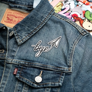 May include: A denim jacket with a white embroidered patch that says "bye" with a paper airplane attached to the word.