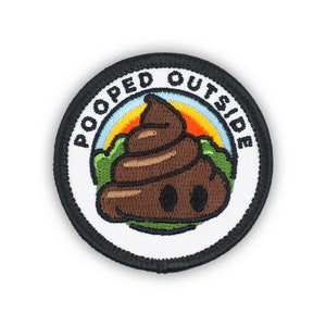 May include: A round, white embroidered patch with black stitching. The patch features a brown cartoon poop emoji with a rainbow and green grass behind it. The text "POOPED OUTSIDE" is written in black around the edge of the patch.