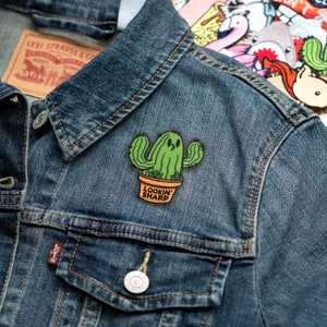May include: A green cactus with a smiling face wearing a pot that says "Lookin' Sharp" is ironed onto a blue denim jacket.