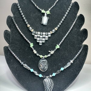 May include: Five necklaces with black beads and various pendants. The pendants include a turtle, a geometric design, a cameo, and a wing. The necklaces are displayed on a black stand.