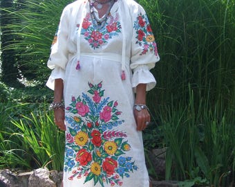 May include: A woman wearing a white linen dress with embroidered floral designs in shades of red, yellow, blue, and green. She is wearing a blue and white headscarf and silver jewelry.
