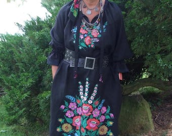 May include: A woman wearing a black embroidered dress with a floral pattern. She is wearing a black head scarf and a silver necklace with multiple strands.