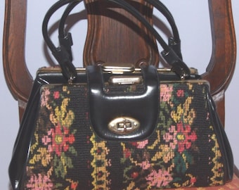 May include: A structured handbag featuring a black base with a multi-colored floral tapestry pattern. The bag includes black handles, a gold-toned metal frame, and a central gold-toned twist-lock closure on a black leather-like tab.