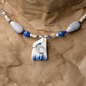 May include: A necklace featuring a central white ceramic pendant with a blue painted base and a silver wire spiral design. The strand consists of grey crackle-patterned beads, blue cylindrical beads, small silver spacers, and light tan wooden cube beads.