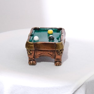 May include: A miniature pool table with a green felt surface and wooden legs. There are three billiard balls on the table: one white, one yellow, and one black.
