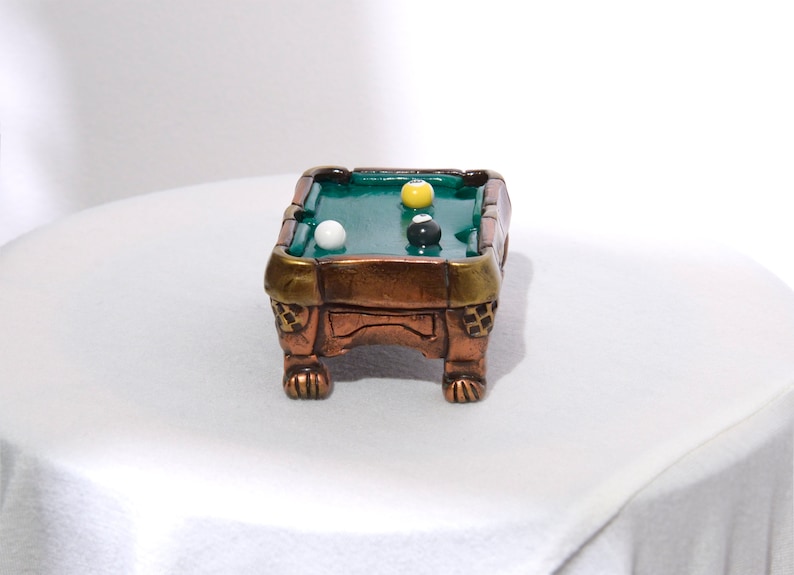 May include: A miniature pool table with a green felt surface and wooden legs. There are three billiard balls on the table: one white, one yellow, and one black.