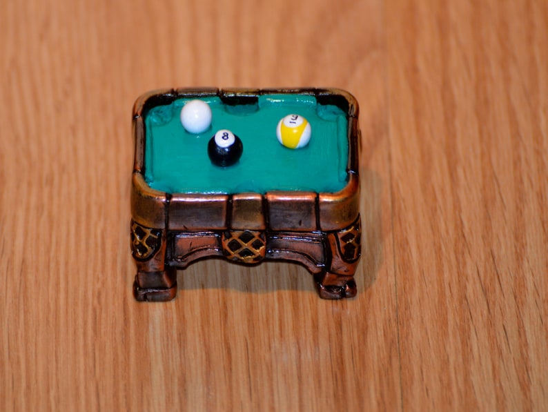 May include: A miniature pool table with a green felt surface and wooden frame. Three billiard balls are on the table: a white ball, a black ball with the number 8, and a yellow ball with black markings.