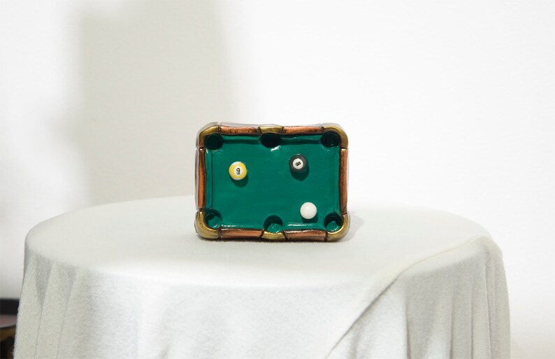 May include: A miniature green felt pool table with a brown wooden frame. The table has three balls on it: a white ball, a black ball with a white number 9, and a yellow ball with a white number 1.