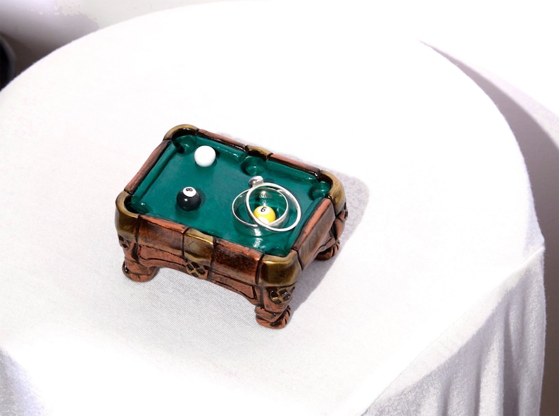 May include: A miniature green felt pool table with a white ball, a black ball with the number 8, and a yellow ball. The table is made of wood with a brown and gold finish.