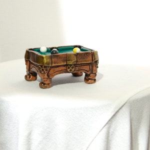 May include: A miniature pool table with a green felt surface and wooden frame. The table has four legs and is decorated with gold accents. There are three billiard balls on the table, one white, one yellow, and one green.