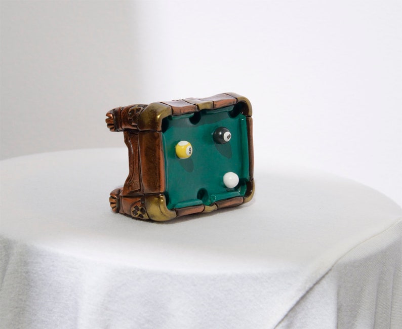 May include: A miniature green felt pool table with brown wooden trim and three billiard balls, one yellow, one black, and one white, on a white surface.