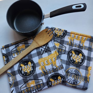 May include: A black saucepan with a black handle, a wooden fork, and two square potholders. The potholders have a black and white checkered pattern with yellow and black text and graphics that read "Fresh Eggs" and "Beef".