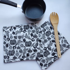 May include: A black saucepan with a black handle, two white and black patterned pot holders, and a wooden cooking spoon. The pot holders feature a repeating pattern of vegetables.