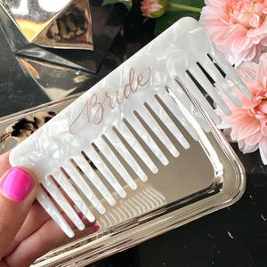 Könnte beinhalten: Ein weißer Kamm mit Perlmutt-Finish, mit dem Wort "Bride" in roségoldener Schrift. Der Kamm liegt auf einem silbernen Tablett, mit rosa Blumen im Hintergrund. Der Kamm wird von einer Person mit pinkfarbenem Nagellack gehalten.