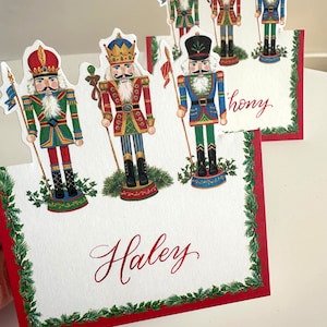 May include: Set of five Christmas place card holders featuring nutcracker illustrations. Each card holder has a red, green, and gold nutcracker with a holly border. The place cards are blank and ready to be written on. The names "Haley" and "Thony" are written on two of the cards.