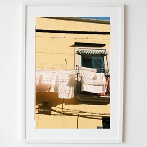 May include: A framed photograph of a yellow building with a window and clothes hanging on a line outside the window.