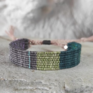 May include: A woven friendship bracelet with a grey, purple, green, and teal color scheme.