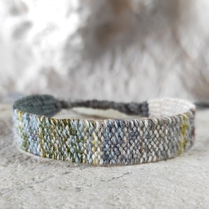 Könnte beinhalten: Gewebtes Armband mit einer Mischung aus Grün, Blau und Grau. Das Armband besteht aus einem strukturierten Material und hat einen verstellbaren Verschluss. Das Armband ist etwa 2 cm breit.