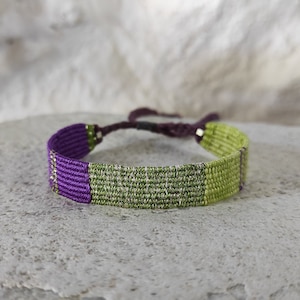May include: A woven bracelet with a green and purple striped pattern. The bracelet is made of thin threads and has a simple design.