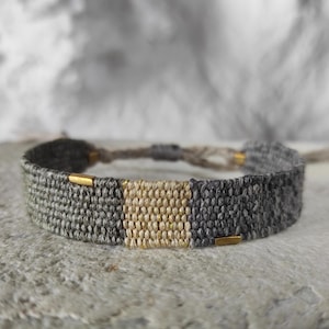 Könnte beinhalten: Ein gewebtes Freundschaftsarmband in Grau und Beige mit goldenen Akzenten.
