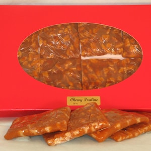 May include: A red box with a clear window showing six pieces of chewy praline candy. The box has the words "Chewy Praline" printed on it. There are also several pieces of praline candy outside of the box.