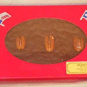May include: A red box with a clear window showing a chocolate pecan candy bar. The box has a gold label that says "HOLIDAY FARMS" and the text "Made in USA".