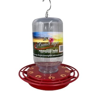 May include: A clear plastic hummingbird feeder with a red base and a metal hook. The feeder has a cylindrical shape with a label featuring a hummingbird and flower design. The base has decorative gold-colored floral patterns.