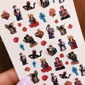 May include: A sheet of nail art stickers featuring images of a couple, a cross, a heart, a rose, and a woman in a religious robe. The stickers are on a white background and have a glossy finish.