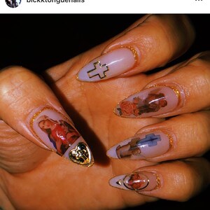 May include: Close-up of a hand with long, almond-shaped nails. Each nail is painted a light pink color and decorated with a gold trim. The nails feature religious imagery, including a cross, a heart, and a figure of a saint.