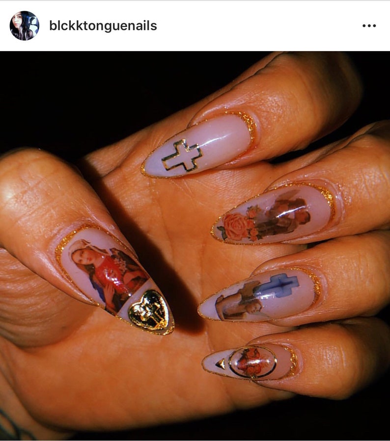 May include: Close-up of a hand with long, almond-shaped nails. Each nail is painted a light pink color and decorated with a gold trim. The nails feature religious imagery, including a cross, a heart, and a figure of a saint.