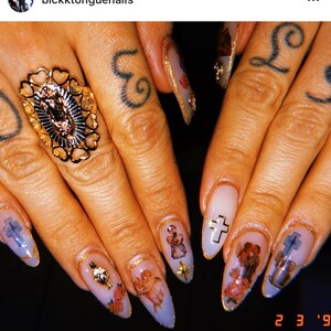May include: Close-up of several long, almond-shaped nails with a light purple base. Each nail is decorated with a gold accent and a different image, including a cherub, a cross, and a religious figure. The nails are on a person's hand, which is wearing a gold ring with a religious image.
