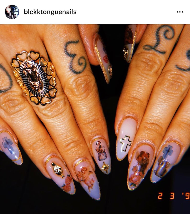 May include: Close-up of several long, almond-shaped nails with a light purple base. Each nail is decorated with a gold accent and a different image, including a cherub, a cross, and a religious figure. The nails are on a person's hand, which is wearing a gold ring with a religious image.