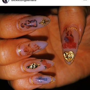 May include: Close-up of long, pointed nails with gold accents and religious imagery. The nails are painted a light pink color and feature images of angels, a cross, and a heart. The nails are decorated with gold glitter and gold trim.