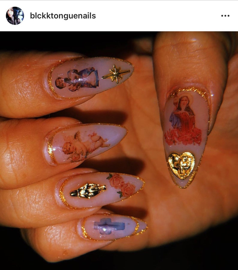 May include: Close-up of long, pointed nails with gold accents and religious imagery. The nails are painted a light pink color and feature images of angels, a cross, and a heart. The nails are decorated with gold glitter and gold trim.