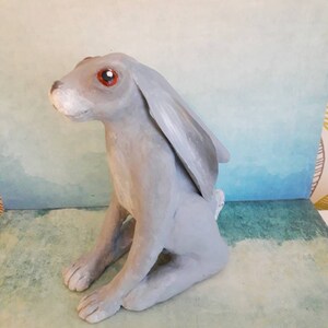 May include: A gray ceramic rabbit sculpture with red eyes. The rabbit is sitting on its hind legs with its front paws resting on its lap. The sculpture is painted in a realistic style.