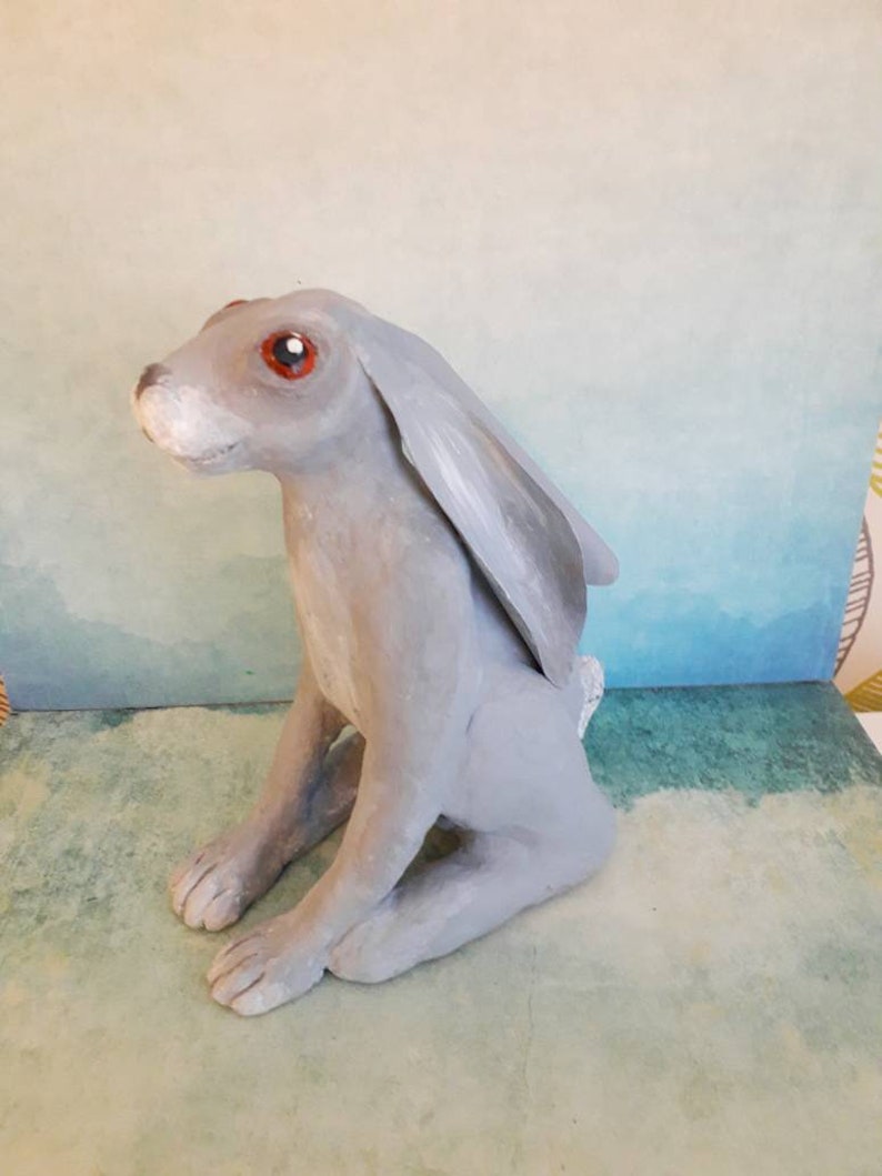 May include: A gray ceramic rabbit sculpture with red eyes. The rabbit is sitting on its hind legs with its front paws resting on its lap. The sculpture is painted in a realistic style.