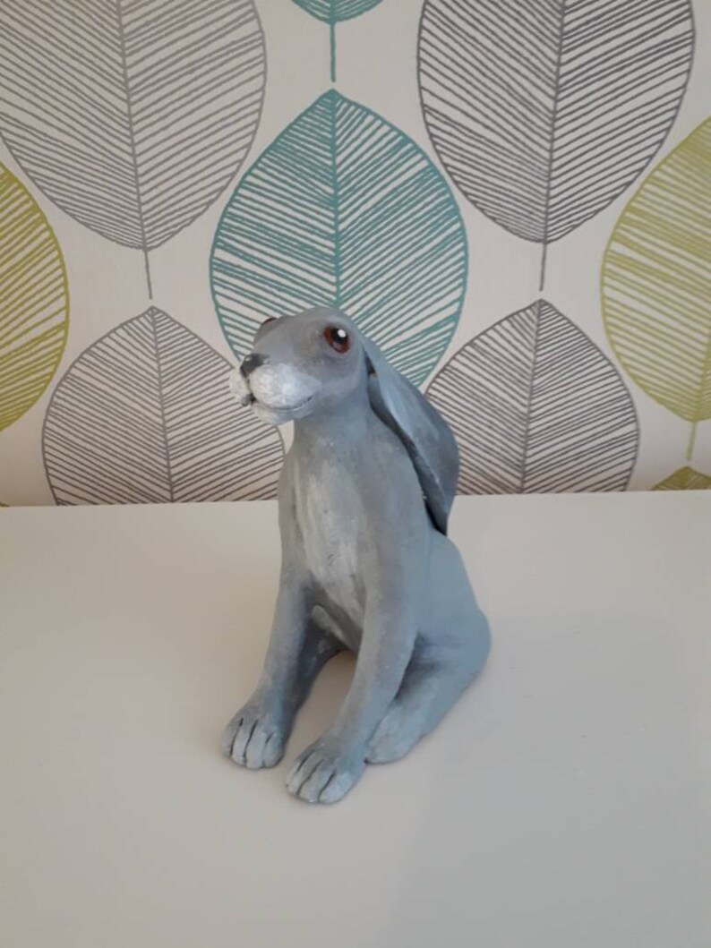 May include: A gray ceramic hare figurine with brown eyes. The hare is sitting with its ears perked up and its tail tucked behind its legs.