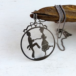 Könnte beinhalten: Eine silberne Halskette mit einem Anhänger, der eine Silhouette eines tanzenden Paares zeigt. Der Anhänger ist aus Metall ausgeschnitten und hat eine runde Form.