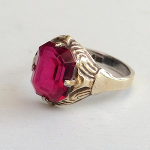 Könnte beinhalten: Ein Vintage-Ring mit einem großen, rechteckigen, rubinroten Edelstein. Der Ring hat eine goldfarbene Fassung mit aufwendigen Details um den Stein und ein silberfarbenes Band. Der Ring ist vor einem weißen Hintergrund platziert.