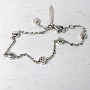 May include: A silver chain bracelet with five round charms, one of which is set with small clear stones. The bracelet has a lobster clasp and a small silver tag with the text "925" on it.