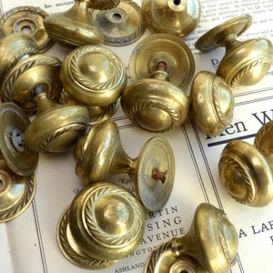 Könnte beinhalten: Eine Sammlung antiker Türknöpfe aus Messing mit verschiedenen Designs, darunter Seil- und Kreisornamente. Die Knöpfe sind auf einem Vintage-Zeitungs-Hintergrund angeordnet, der ihre gealterte Patina und ihren klassischen Stil zeigt.