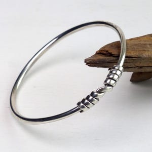 Könnte beinhalten: Ein silbernes Armreif-Armband mit einem gedrehten und gewickelten Design. Das Armband besteht aus glänzendem, poliertem Metall und hat einen schlichten, eleganten Stil. Ein perfektes Accessoire für den Alltag.