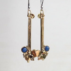 May include: A pair of long, vertical gold-toned metal earrings featuring a smooth rectangular bar suspended from a hook. The base of each earring is adorned with a cluster of small metallic spheres, flat circular discs, and a single round blue stone.