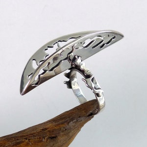 May include: A silver ring with a unique design. The top of the ring is a half-circle with a cut-out pattern. The band of the ring is also intricately designed.