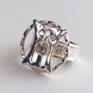 Puede incluir: Un anillo de plata que presenta un marco circular con una figura abstracta y estilizada similar a un animal en el centro. La figura incluye un pequeño elemento de anillo colgante. La banda del anillo tiene un acabado de superficie texturizado.